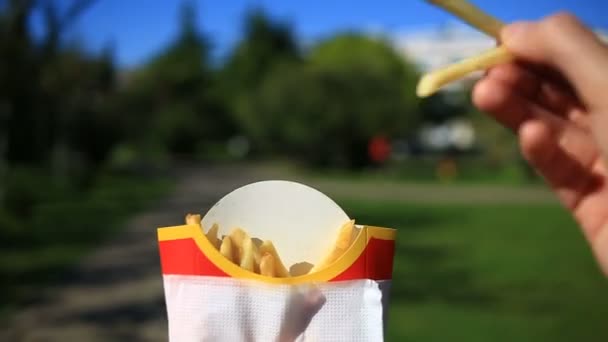 En man äter snabbmat på gatan. Han bär en pommes frites och äter. Mot bakgrund av en suddig stadsgata — Stockvideo