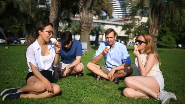 Amigos sentar em uma clareira verde no meio da cidade e conversar, eles comem sorvete em um dia quente de verão — Vídeo de Stock