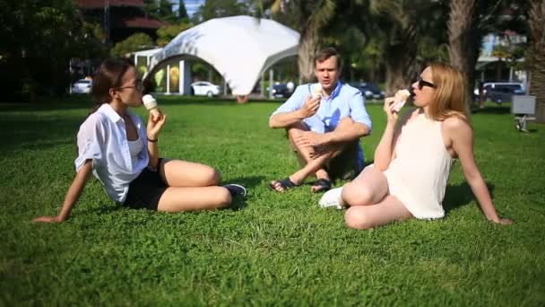 Amigos se sientan en un claro verde en el centro de la ciudad y hablan, comen helado en un día caluroso de verano — Vídeos de Stock