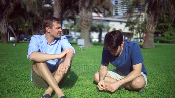 Dos amigos están sentados en un claro verde en medio de la ciudad y hablando, los homosexuales están descansando — Vídeos de Stock