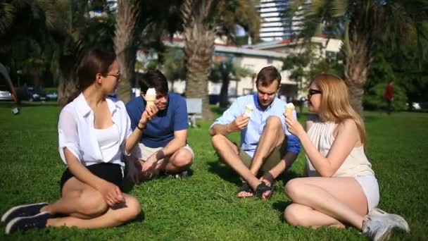 Друзі сидять у зеленій галявині посеред міста і розмовляють, вони їдять морозиво в спекотний літній день — стокове відео