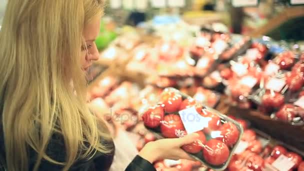 Μια γυναίκα σε ένα σούπερ μάρκετ σε ένα ράφι λαχανικών, αγοράζει τα λαχανικά και τα φρούτα. Ο άνθρωπος επιλέγει μια ντομάτα — Αρχείο Βίντεο