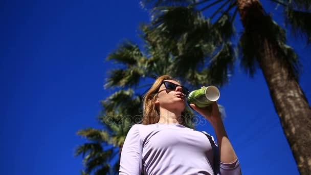 La jeune fille boit un verre dans la rue à partir d'une tasse jetable contre le ciel bleu et les palmiers. Vue du bas — Video
