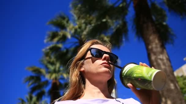 Das Mädchen trinkt auf der Straße ein Getränk aus einem Einwegbecher vor blauem Himmel und Palmen. von unten — Stockvideo