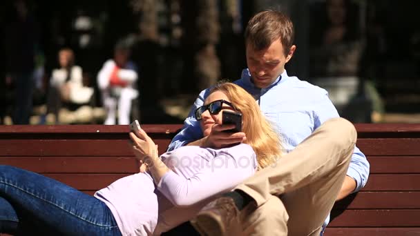 Feliz jovem casal relaxante no parque em um banco, menina inclinada no colo de seu namorado e olhando para o telefone — Vídeo de Stock