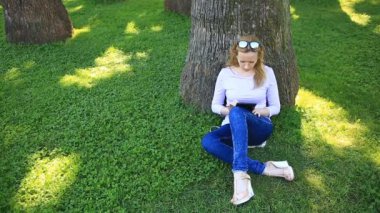 Bir palmiye ağacının altında oturan ve browsing belgili tanımlık enterne üstünde bir smartphone yakışıklı kadın