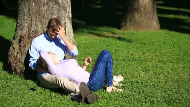 Pareja romántica sentada bajo una palmera. Una chica en el regazo de un chico. Una pareja amorosa descansando en un parque en la hierba bajo un árbol — Vídeos de Stock