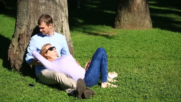 Ρομαντικό ζευγάρι κάθεται κάτω από ένα φοίνικα. Ένα κορίτσι στην αγκαλιά ενός άντρα. Ένα ζευγάρι ερωτευμένων που στηρίζεται σε ένα πάρκο στο γρασίδι κάτω από ένα δέντρο — Αρχείο Βίντεο