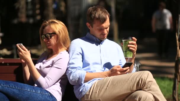 Hombre y mujer mirando en diferentes direcciones, sentados en un banco. Todo el mundo está mirando su teléfono móvil — Vídeos de Stock