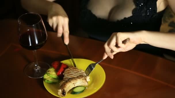 Un primo piano di una donna con una scollatura taglia un pezzo di costolette in ossa cotte su una griglia. Donna sexy mangiare carne e bere vino — Video Stock