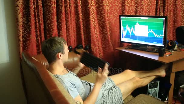 A man in a T-shirt and shorts, sitting at home on the couch, monitors changes in the schedule on the currency exchange, looking at the computer monitor — Stock Video