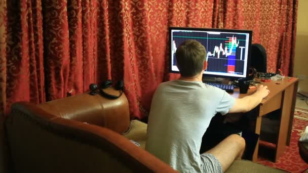A man in a T-shirt and shorts, sitting at home on the couch, monitors changes in the schedule on the currency exchange, looking at the computer monitor