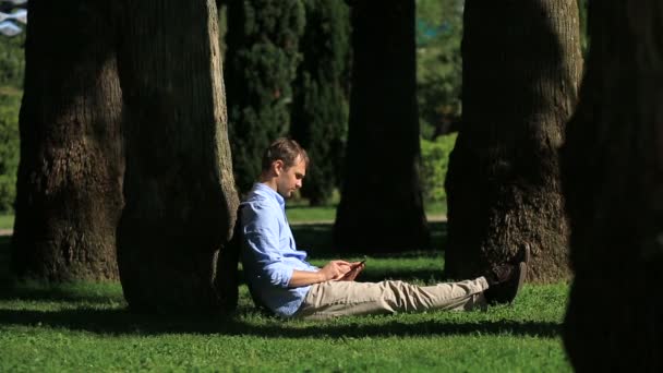 Όμορφος άνδρας κάθεται κάτω από ένα φοίνικα και περιήγηση στο Internet σε ένα smartphone — Αρχείο Βίντεο