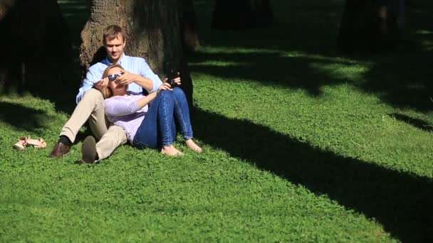 Pareja romántica sentada bajo una palmera. Una chica en el regazo de un chico. Una pareja amorosa descansando en un parque en la hierba bajo un árbol — Vídeo de stock