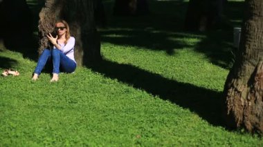 Bir palmiye ağacının altında oturan ve browsing belgili tanımlık enterne üstünde bir smartphone yakışıklı kadın
