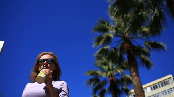 A lány az utcán egy eldobható pohár ellen a kék ég- és pálmafák között iszik egy italt. Alulnézet — Stock videók