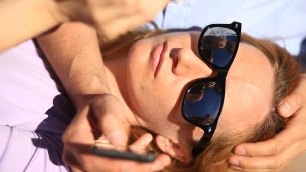 Happy young couple relaxing in the park on a bench, girl leaning on the lap of her boyfriend and looking at the phone — Stock Video