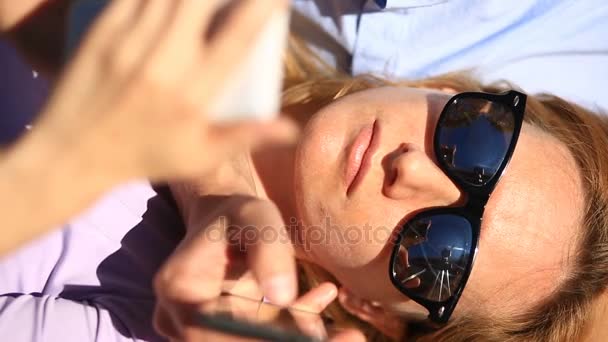 Feliz jovem casal relaxante no parque em um banco, menina inclinada no colo de seu namorado e olhando para o telefone — Vídeo de Stock