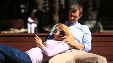 Mutlu genç çift kız sevgilisinin kucağına yaslanmış ve seyir telefon parkta bir bankta rahatlatıcı