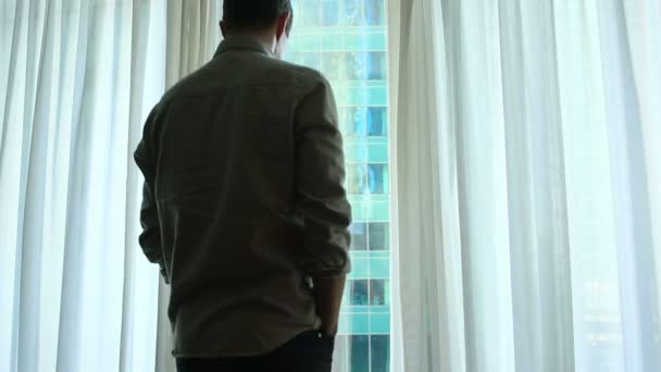 A man stands by the window with curtains in a hotel room, looks at an amazing city landscape — Stock Video