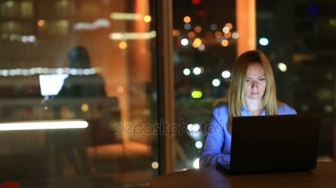 Güzel sarışın iş kadını fazla mesai çalışma gece çift ofisinde. Şehir ışıkları arka plan büyük bir pencere görünür