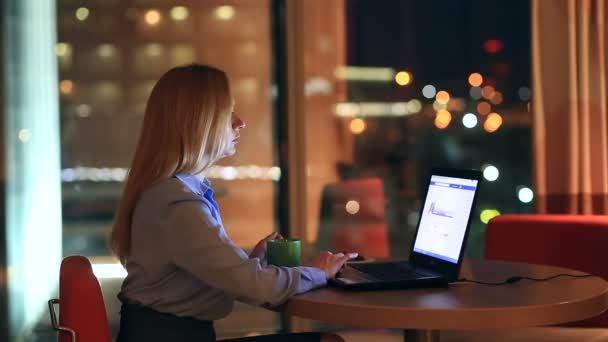 Hermosa mujer rubia de negocios trabajando horas extras por la noche en la oficina ejecutiva. Las luces de la ciudad son visibles en el fondo desde una ventana grande — Vídeo de stock