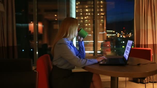 Hermosa mujer rubia de negocios trabajando horas extras por la noche en la oficina ejecutiva. Las luces de la ciudad son visibles en el fondo desde una ventana grande — Vídeo de stock