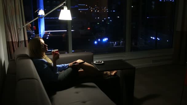 Žena leží na pohovce s hrnkem v ruce u okna s noční pohled na město. sledování Tv — Stock video