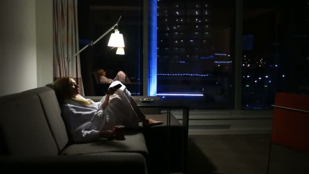 Une femme est allongée sur un canapé avec une tasse dans les mains près de la fenêtre avec une vue de nuit sur la ville. regarder la télévision — Video
