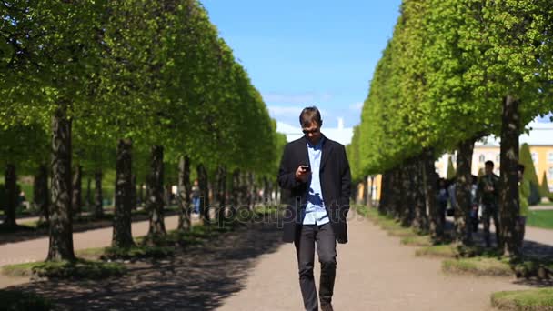 Ein Mann mit Handy geht langsam an einem grünen Park vorbei, ein Mann hält ein Handy in der Hand, — Stockvideo