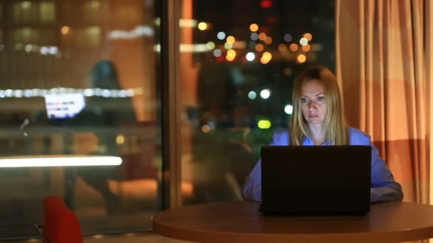 美しい金髪のビジネスの女性は、事務局で、夜残業します。街の灯が大きな窓からバック グラウンドで表示されます。 — ストック動画