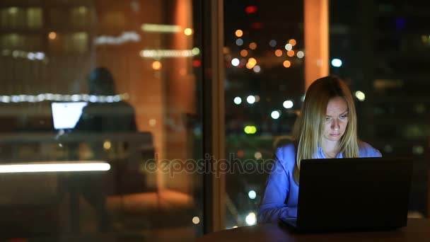 Hermosa mujer rubia de negocios trabajando horas extras por la noche en la oficina ejecutiva. Las luces de la ciudad son visibles en el fondo desde una ventana grande — Vídeos de Stock