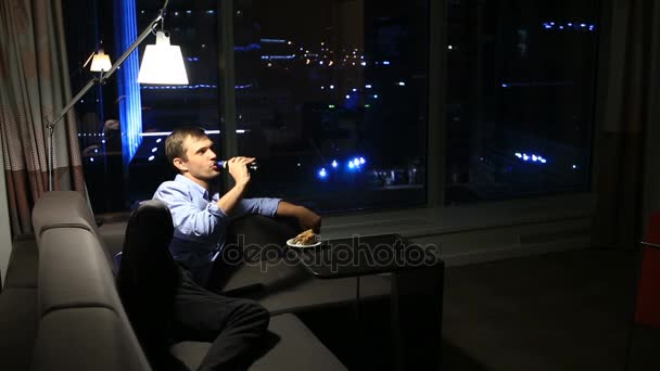 Мужчина отдыхает дома на диване ночью, пьет пиво и смотрит телевизор, на фоне большого понамрамного окна с видом на ночной город — стоковое видео