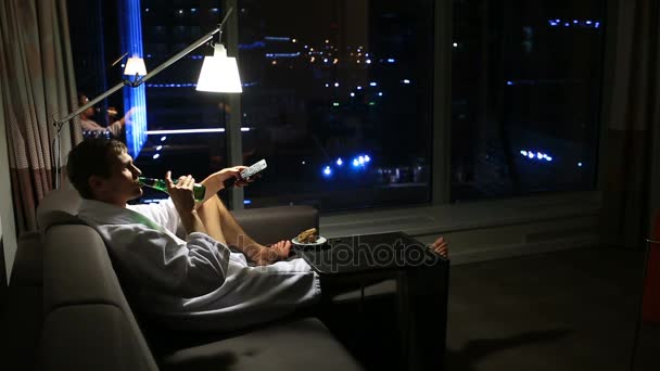 L'homme se repose à la maison sur le canapé la nuit, boire de la bière et regarder la télévision, sur le fond d'une grande fenêtre ponamramnogo donnant sur la ville de la nuit — Video