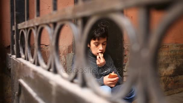Безпритульний підліток їсть кору хліба у ворота на тлі решітки — стокове відео