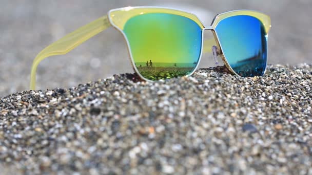 Zonnebril op het zand, het strand met de rust mensen wordt weerspiegeld in de glazen. Gouden bril — Stockvideo