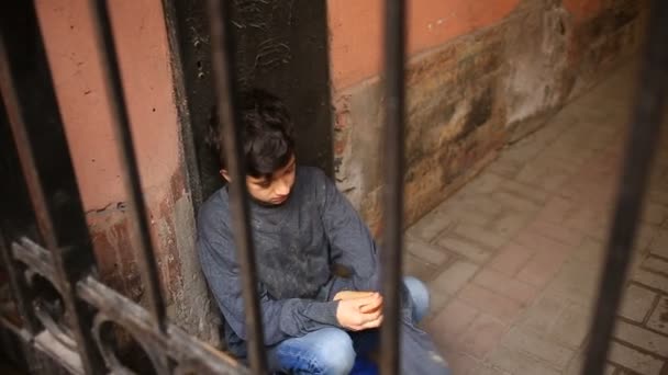 Бездомный подросток ест корочку хлеба в воротах на фоне решетки — стоковое видео