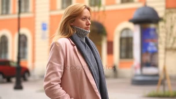 Uma mulher cansada em um casaco rosa e suéter fica no meio de uma rua lotada e encolhe do frio — Vídeo de Stock