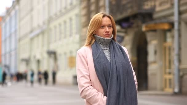 Una mujer cansada con un abrigo rosa y suéter se encuentra en medio de una calle llena de gente y se encoge del frío — Vídeos de Stock