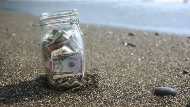 Un frasco de vidrio con dólares de papel moneda contra un fondo de olas marinas. Acumular los ahorros en vacaciones por el mar o el océano. playa — Vídeos de Stock