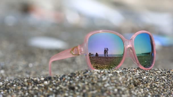 Óculos de sol na areia, a praia com as pessoas em repouso é refletida nos óculos. Óculos rosa — Vídeo de Stock