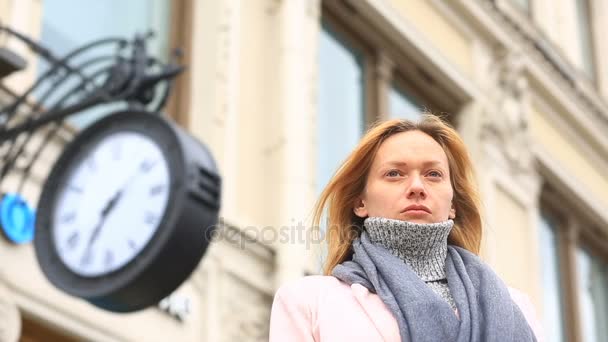 Женщина в розовом пальто и свитере стоит посреди переполненной улицы рядом с часами и ждет встречи — стоковое видео