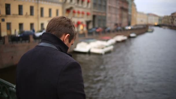 Un hombre con un abrigo se para en un puente y mira hacia el agua negra. Quiere suicidarse. — Vídeo de stock