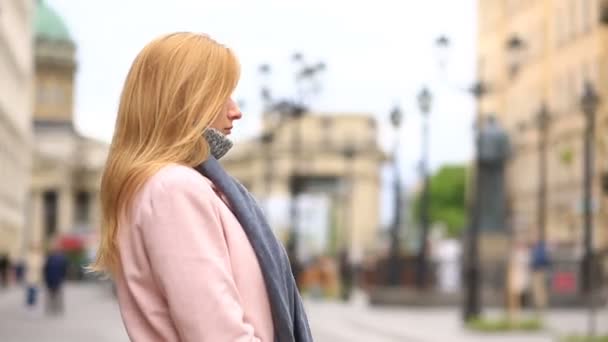 Камера ковзає навколо жінки, яка стоїть посеред міської вулиці в рожевому пальто і шарфі — стокове відео