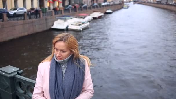 Vrouw in roze jas en trui staat op brug over water achtergrond en kijkt naar de camera — Stockvideo