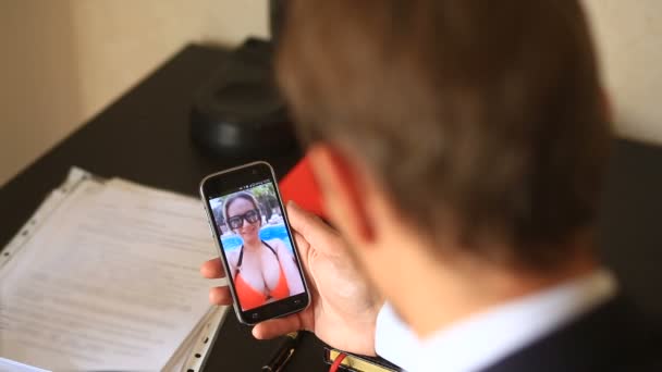 Un hombre de negocios con traje, sentado en la oficina de su escritorio, mantiene un video chat con una chica en la playa. Ella está vestida con un traje de baño y lo invita a unirse a ella — Vídeo de stock