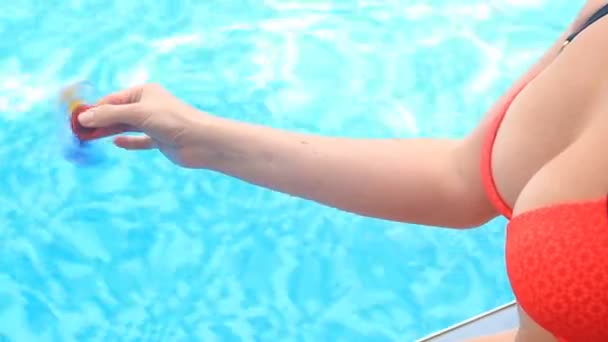 Un spinner o spinner multicolor, rojo-amarillo-azul de la mano, girando en el brazo de las personas. Una mujer haciendo girar un Fidget Spinner sobre un fondo de agua azul en una piscina cerca de sus exuberantes pechos en bikini — Vídeos de Stock