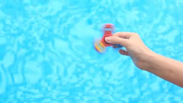 Un spinner o spinner multicolor, rojo-amarillo-azul de la mano, girando en el brazo de las personas. Una mujer haciendo girar un Fidget Spinner sobre un fondo de agua azul en una piscina — Vídeo de stock