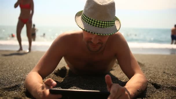 男人在海滩上使用电子的平板电脑。戴着帽子的男人躺在海滩上，看着平板电脑 — 图库视频影像