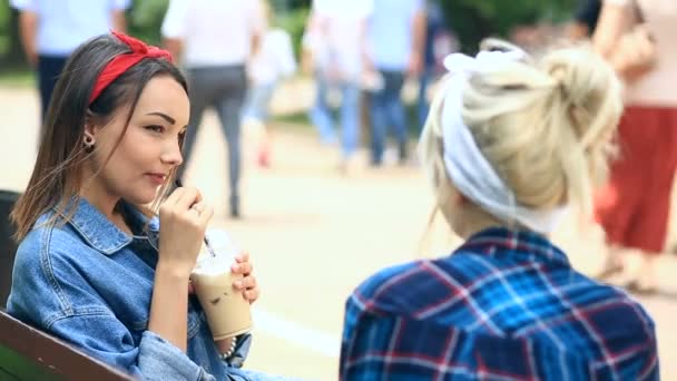 Δύο φίλοι κάθονται σε ένα παγκάκι στο πάρκο της πόλης και πιείτε ένα milkshake από ένα φλιτζάνι μιας χρήσης — Αρχείο Βίντεο